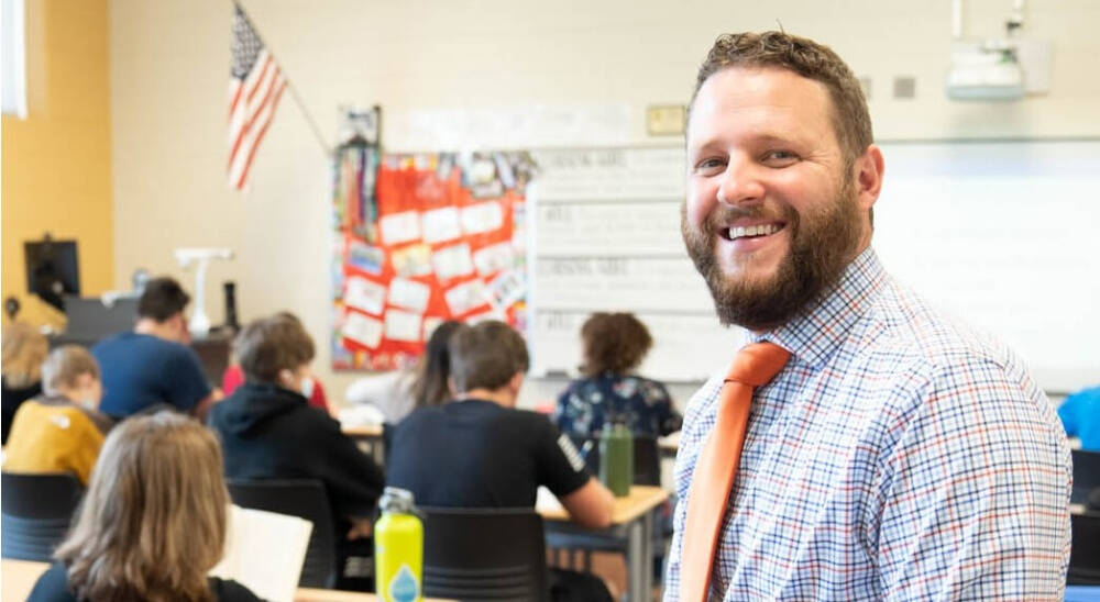 A smiling person in a classroom, wearing a checkered shirt and an orange tie, with students seated in the background.